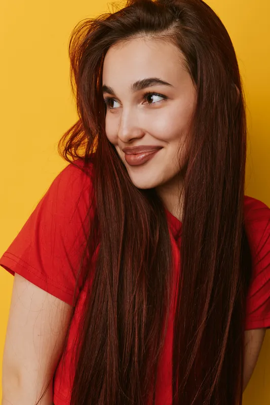 Young woman with long red hair wearing red shirt smiling and looking sideways against yellow background. Portrait of happy female with natural makeup and casual style indoors.