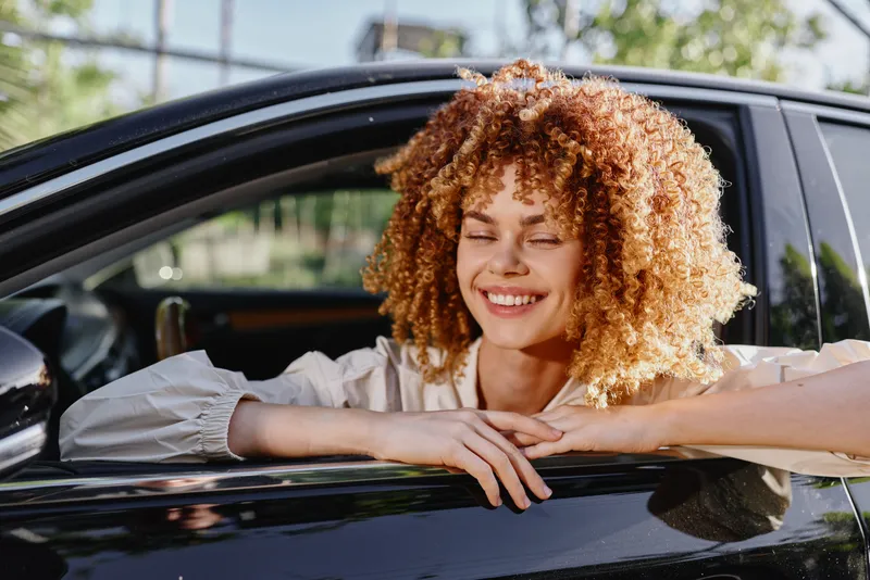 Smiling woman with curly hair leaning out of car window on sunny day, wearing light blouse, enjoying fresh air and relaxed moment outdoors in casual style.