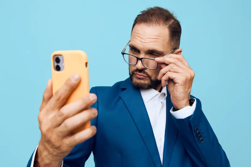 A professional man in a blue suit stands against a solid blue background, focused on a smartphone with a thoughtful expression, conveying business insight and curiosity.
