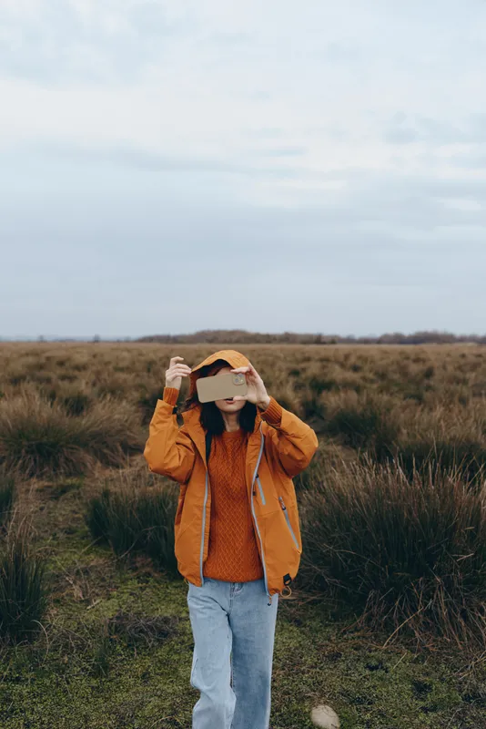 Woman outdoors in grassy field lifestyle, wearing a warm hat and holding it up, shielding eyes, relaxed standing pose, soft sunlight, natural colors, open sky, in spring air, quietly