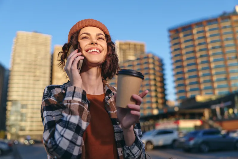 Woman with coffee and phone in city morning, smile and beanie in an urban scene. Candid moment with authenticity, golden hour glow, mindful living and emotional storytelling vibe.