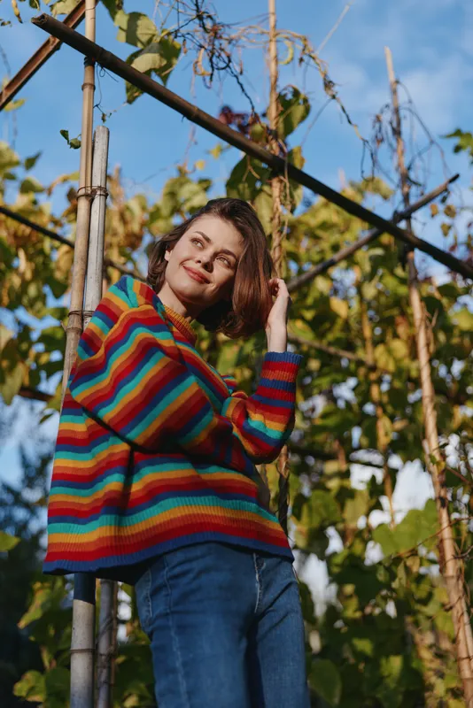 Woman in a colorful striped sweater and casual outfit with jeans, smiling and leaning on a bamboo trellis in a sunny garden. Young adult portrait with relaxed outdoor vibe.