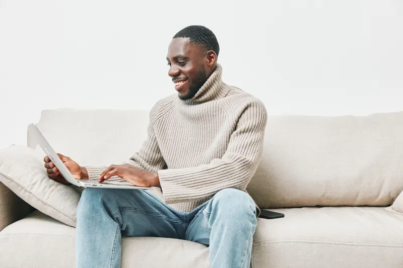 Young African man smiling while working on a laptop at home, wearing a cozy beige sweater, seated on a light colored couch, exuding a relaxed and productive atmosphere