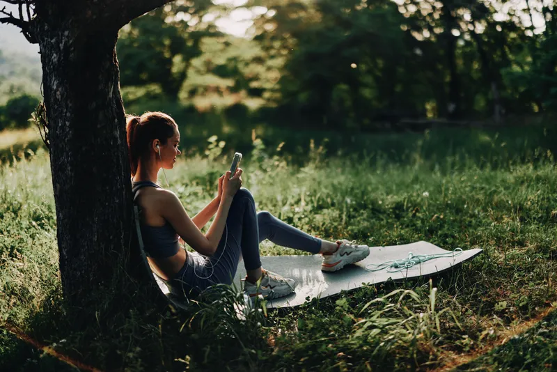 Woman in athletic wear relaxing outdoors, enjoying nature while using a smartphone, surrounded by greenery Healthy lifestyle and mindfulness concept