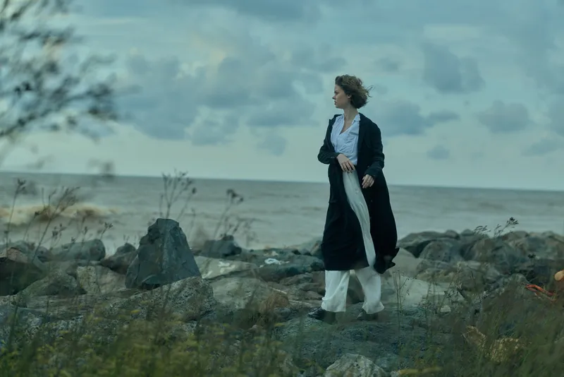 Woman walking along a rocky coast by the sea on a windy day, wearing a long cardigan and flowing dress, with a moody sky and open horizon in view of distant waves rolling to shore