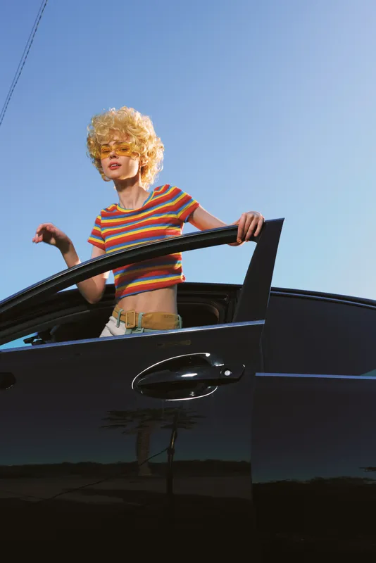 A stylish woman wearing a blonde curly wig leans from a glossy car window, wearing a striped top. Retro fashion vibe highlights bold confidence and playful street style.