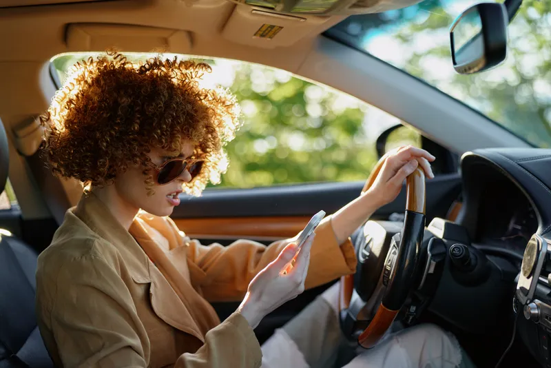A woman driver in a tan jacket checks her phone while seated in a sunlit car, capturing a relaxed lifestyle moment with retro film color and casual fashion, urban vibe.
