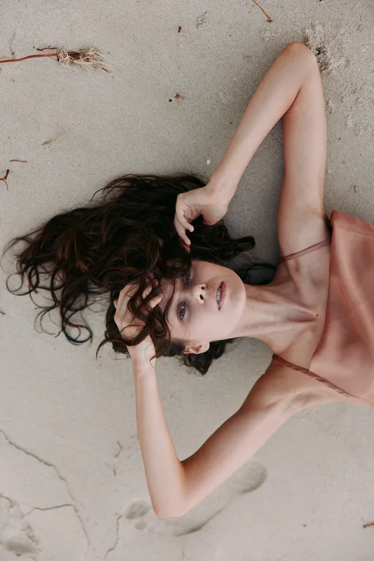 A graceful woman lies on sandy beach in a soft pink dress, sunlit skin and dark hair forming gentle lines, capturing a serene, timeless moment.