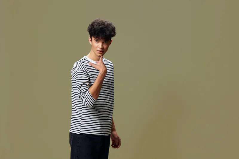 young man in striped shirt with curly hair standing against plain beige background, posing casually with a thoughtful expression, minimalistic studio setup