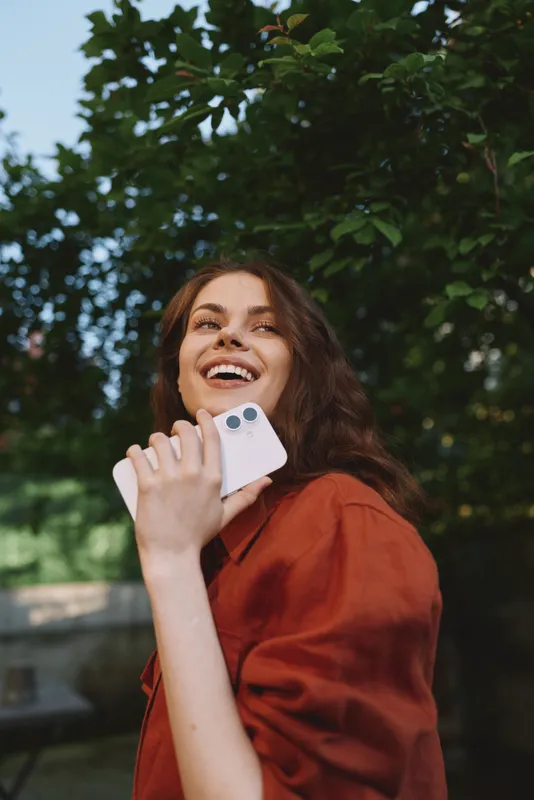 Smiling woman outdoors in a red shirt, holding a smartphone. Natural light, lush greenery, candid lifestyle moment, warm, cheerful, relaxed vibe