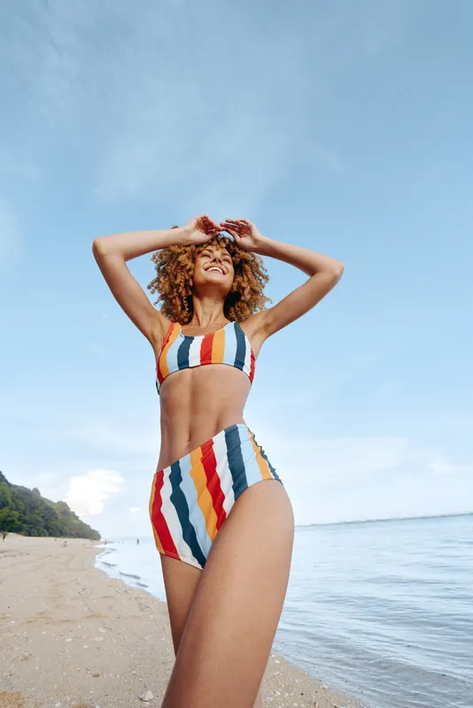 Woman standing on beach by ocean, colorful swimsuit with stripes, summer sunlight, outdoor seaside setting, arms behind head pose, natural light, carefree lifestyle mood, waves and sky blue