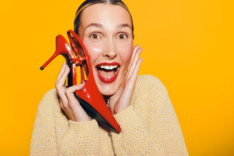 Excited woman in yellow sweater holds red high heel shoe near face with open mouth on bright yellow background. Concept of fashion, style, happiness, and shopping joy.