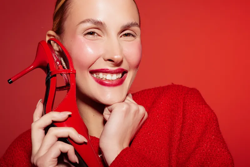 Smiling woman in red sweater holds red high heel shoe near face, showing bright makeup and joyful expression. Concept of fashion, beauty, and happiness on red background.