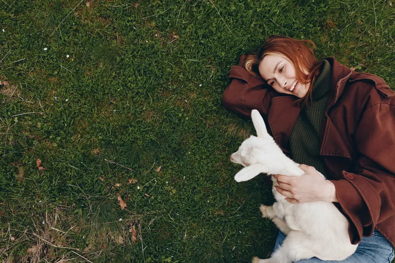 Relaxed woman lies on grass cuddling a white dog outdoors, wearing a brown coat, serene mood, candid lifestyle moment, warm natural light, simple affectionate scene