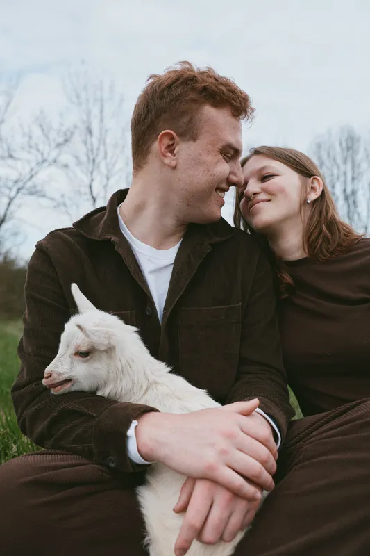 A couple outdoors shares a tender moment with a playful goat kid, smiling at each other while embracing in a rural setting. A warm lifestyle scene highlighting connection and affection.