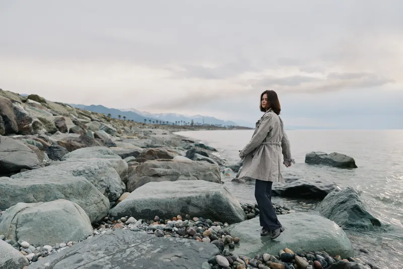 A young woman stands on rocky shore near calm sea under cloudy sky. She wears casual clothes and looks over her shoulder. Concept of nature, solitude, and outdoor lifestyle.