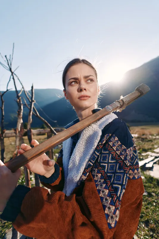 Woman outdoors in mountains holding a wooden staff across her shoulders, rural portrait at sunrise with patterned jacket and meadow valley background, thoughtful gaze in warm sunlight.