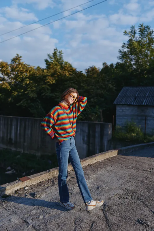 Woman in striped sweater and blue jeans standing outdoors on a rooftop, smiling and relaxed, early twenties model with casual outfit, hand in hair, lifestyle fashion travel vibe.