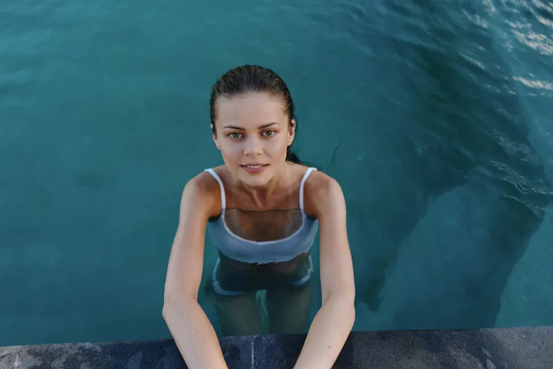 Smiling woman in a white swimsuit emerges from a blue pool, hands gripping the edge. Close-up of wellness, fitness, and lifestyle with water reflections and warm sunlight creating mood.