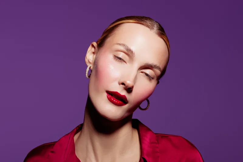 Portrait of a woman with closed eyes wearing red lipstick and earrings on purple background. Concept of beauty, makeup, fashion, and elegance in studio lighting.