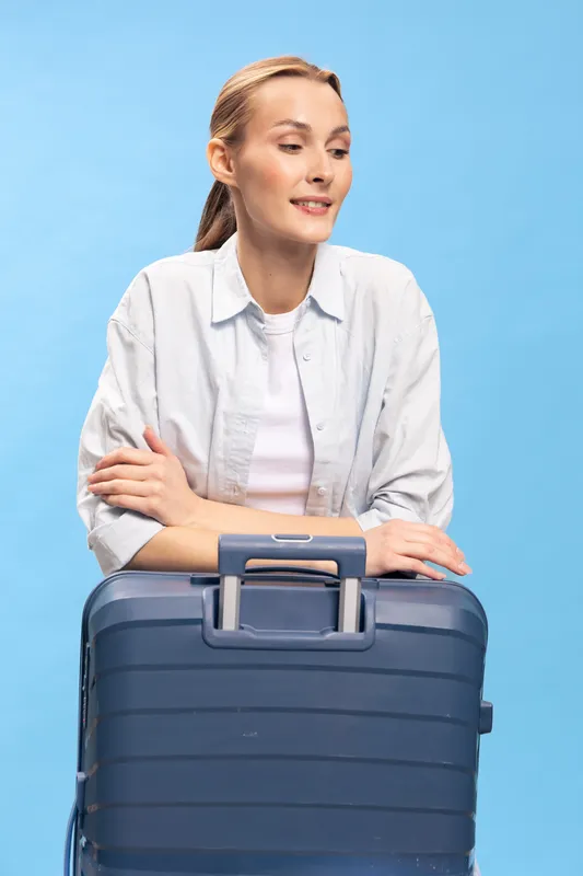 Young woman with light hair leans on blue suitcase handles, looking sideways with a slight smile against a blue background. Concept of travel, vacation, and leisure time.