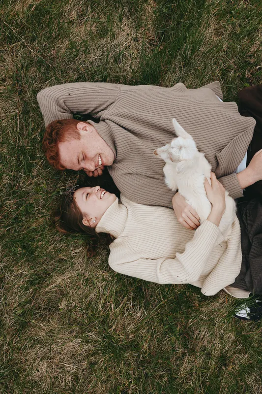 Loving couple lying on grass cuddling a small goat kid under soft daylight, outdoors lifestyle scene, affection, nature, warmth, serenity, happiness, tenderness, connection, calm, relaxed mood.