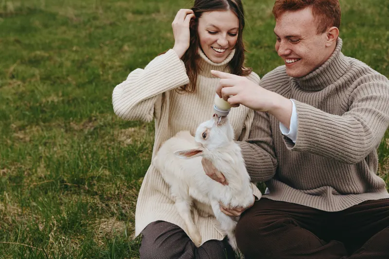 Smiling couple feeding a baby goat with a bottle outdoors on green grass. Cozy sweaters, soft natural light, rustic mood. Lifestyle moment focused on care, bonding, and farm animals.