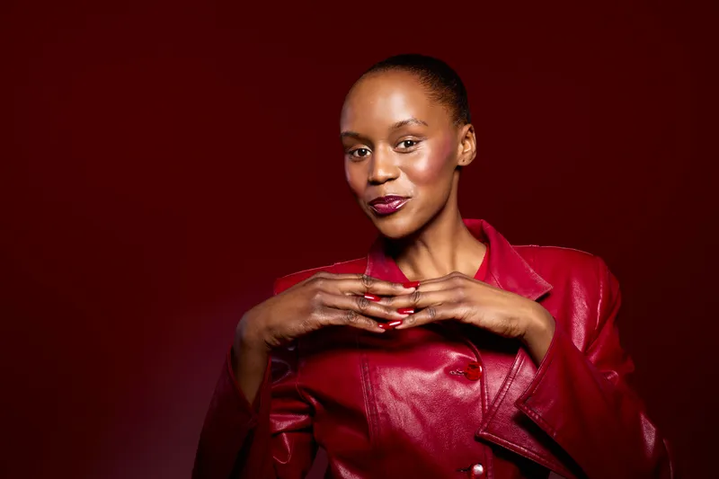 Confident African woman with dark skin wearing glossy red leather jacket poses against red background. Studio portrait of stylish female model with natural makeup and elegant expression.