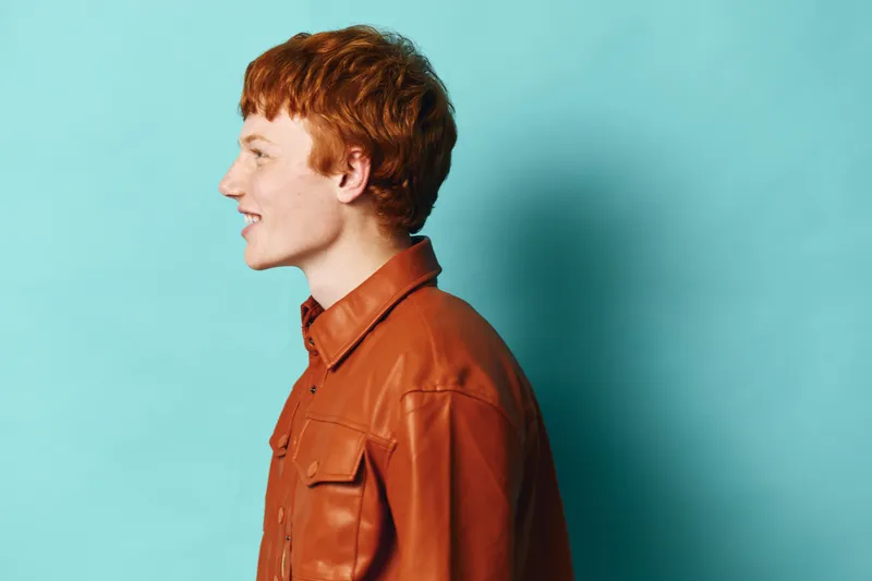 Profile of young man with red hair and freckles wearing brown leather jacket against blue background. Casual portrait with relaxed expression and short hairstyle in studio lighting.