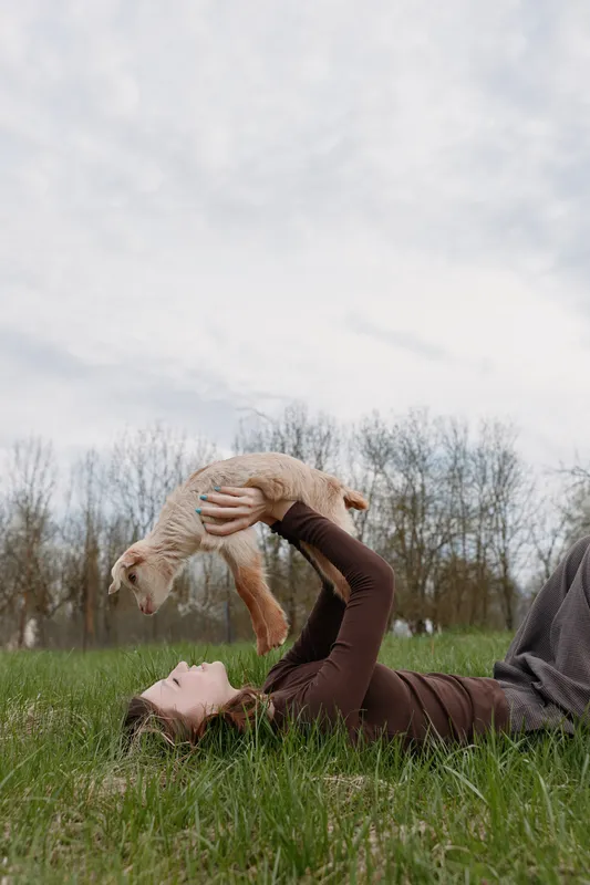 Young woman lies on green grass holding a baby goat above her with joy in a natural outdoor setting under cloudy sky
