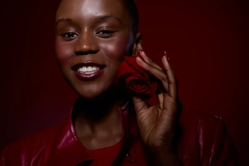 Smiling black woman with dark skin holds red rose near face wearing red leather jacket. Studio portrait with dark red background. Concept of beauty, love, and confidence for people of color.