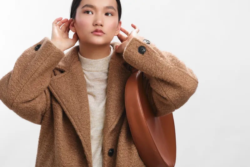 an asian woman wearing a warm tan coat and soft knit sweater adjusts her earpiece in a bright studio, presenting a calm, confident, and stylish attitude.