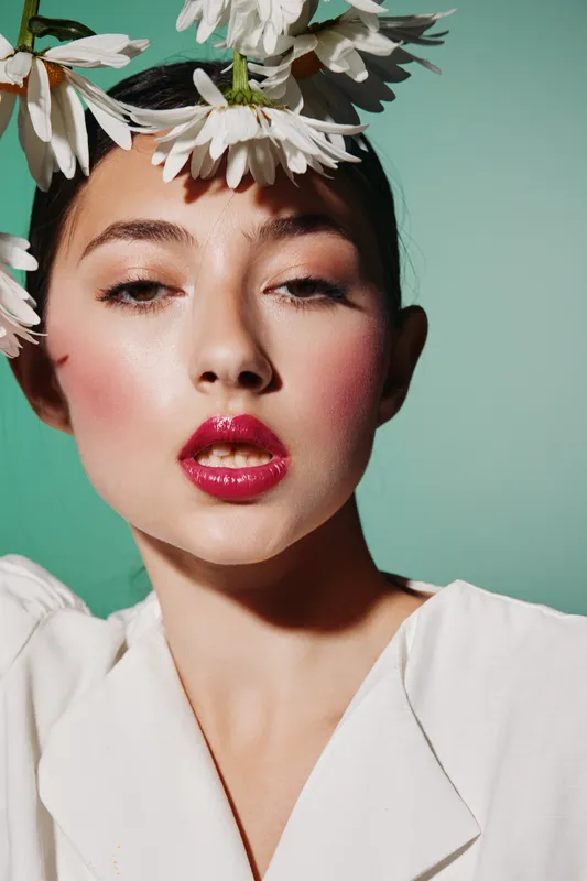 Close-up of a young woman with bright red glossy lips and rosy cheeks surrounded by white flowers against a turquoise background. Concept of beauty, fashion, creativity, makeup.