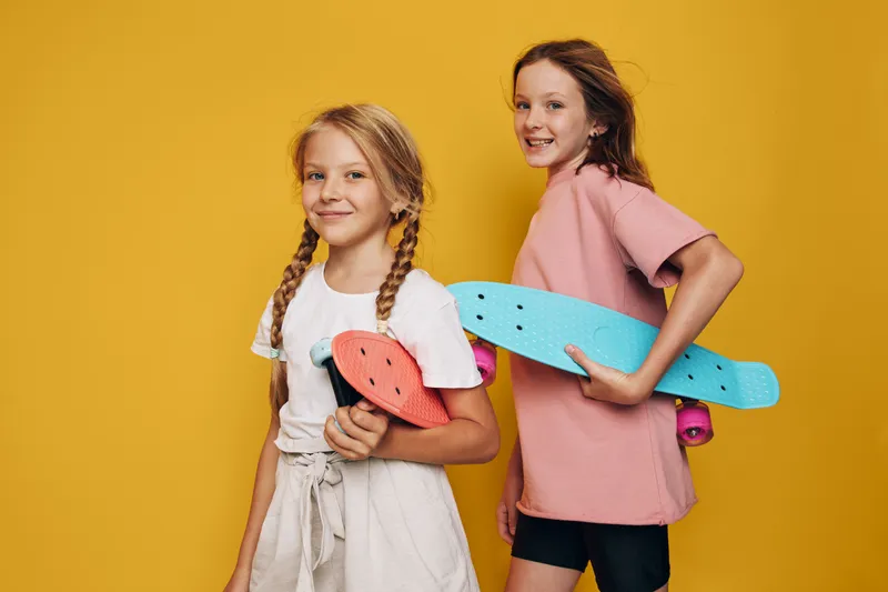 Two smiling girls holding colorful skateboards stand against a vibrant yellow background. Both wear casual summer clothes and look confident and happy. Youthful fun concept.