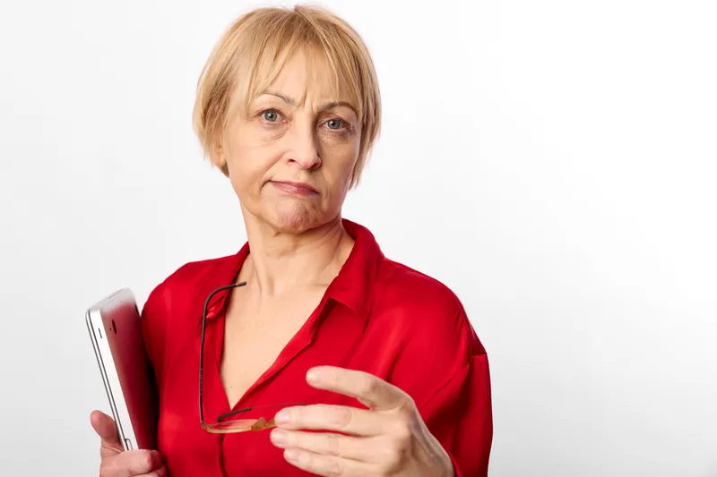 Senior woman stressed in red medical uniform holds glasses, worried expression, healthcare clinic concept for anxiety, concern and mental health, white background portrait
