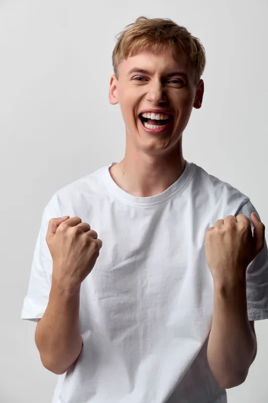 Cheering, excited man in a white t shirt shows triumph with clenched fists and a wide smile, conveying energy, happiness and celebration after a win or achievement