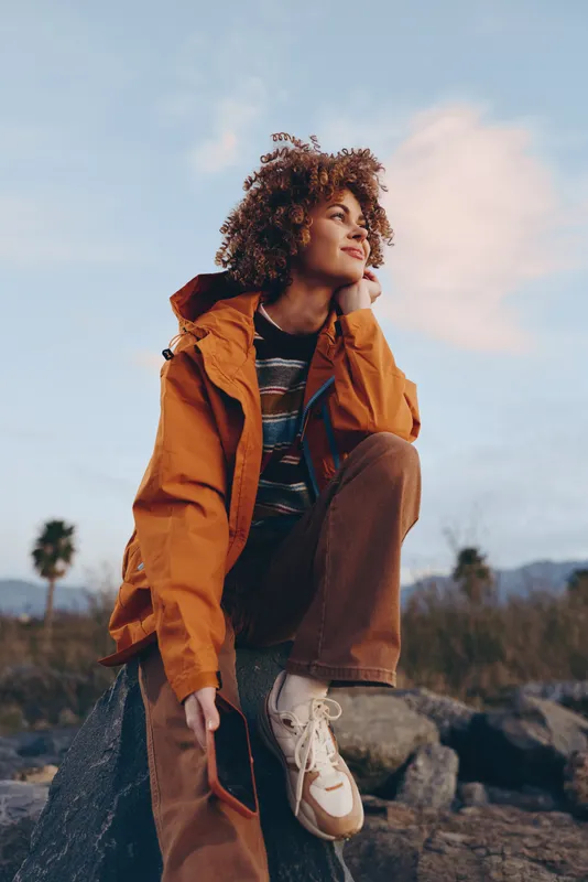 Curly haired woman outdoors, wearing orange jacket, smiling softly, relaxed lifestyle pose on rocky ground, blue sky, distant palms, natural light