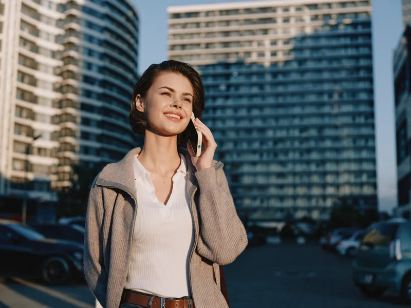 A confident businesswoman stands outdoors in a modern city, speaking on a smartphone. She wears smart casual attire, daylight highlights her smile, projecting professionalism and a dynamic, energetic