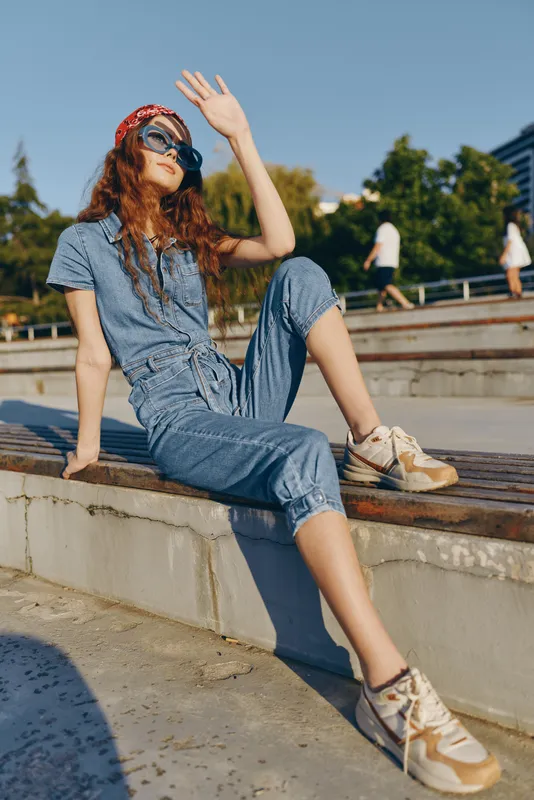 relaxed woman in denim jumpsuit sitting outdoors on concrete steps in sunny weather with sunglasses and red headband enjoying warm summer day