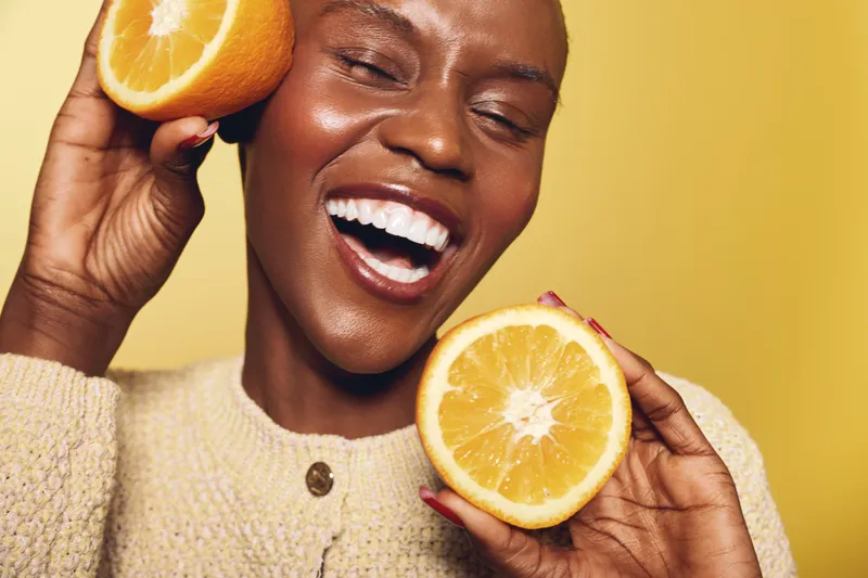 Happy African woman with dark skin smiling widely holds two halves of fresh orange fruit near her face on yellow background. Concept of healthy lifestyle, vitamin C, and natural beauty.
