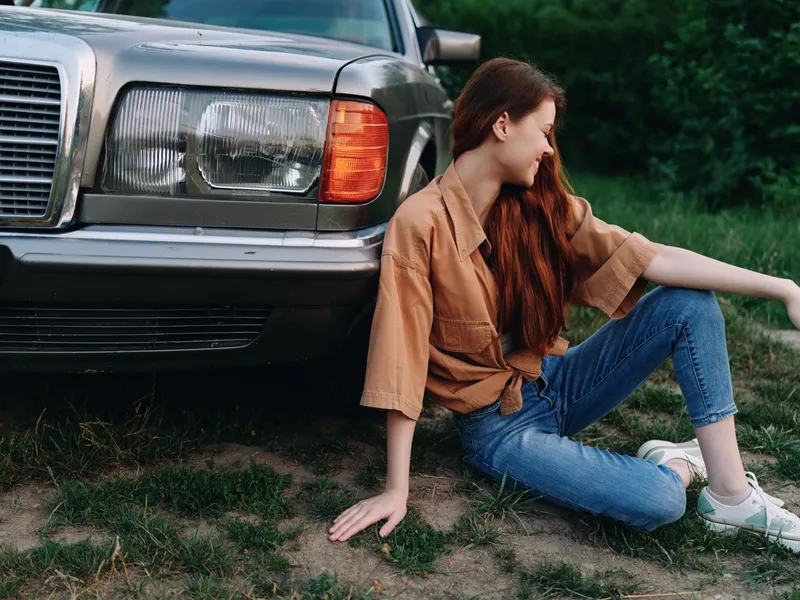 Young woman with long hair in casual outfit sitting by an old car, enjoying a warm day outdoors, exuding relaxation and happiness in a natural setting.