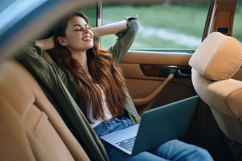 Relaxed young woman in car using laptop, smiling with sunlight, showcasing remote work lifestyle, enjoying calmness and comfort in nature.
