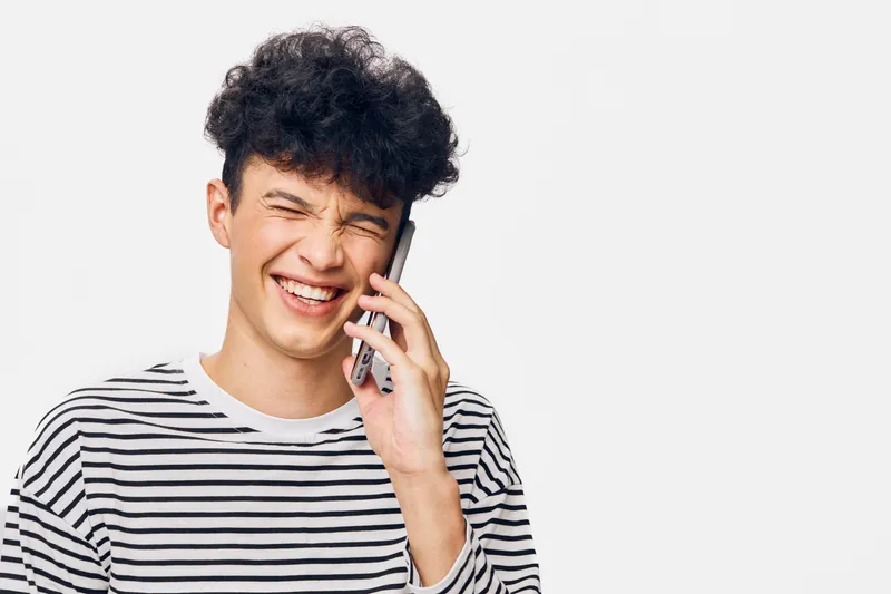 Smiling man wearing striped shirt talks on phone with joyful expression, curly hair, and bright mood against white background, casual and lively communication moment.