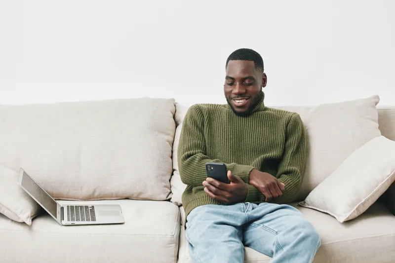 Joyful young Black man in cozy green sweater smiling while using smartphone on beige couch with laptop nearby, enjoying leisure time at home
