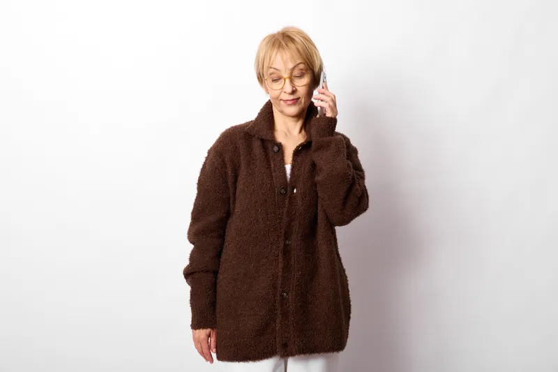 Elderly woman holding phone to ear in a studio portrait, cozy brown coat, calm focused expression, neutral light background, soft even lighting, quiet indoor atmosphere