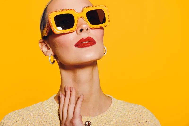 Stylish woman with short hair wearing orange sunglasses and red lipstick poses against bright yellow background. Fashion portrait with confident expression and modern accessories.