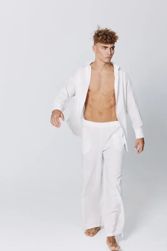 Young man with curly hair and bare chest dressed in white loose pants and open shirt walking barefoot on light studio background with confident expression and casual style.