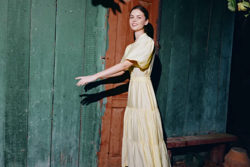 Young woman in a pale yellow dress stands beside a weathered wooden wall posing with a gentle smile capturing a bright outdoor portrait with soft light and calm vibe