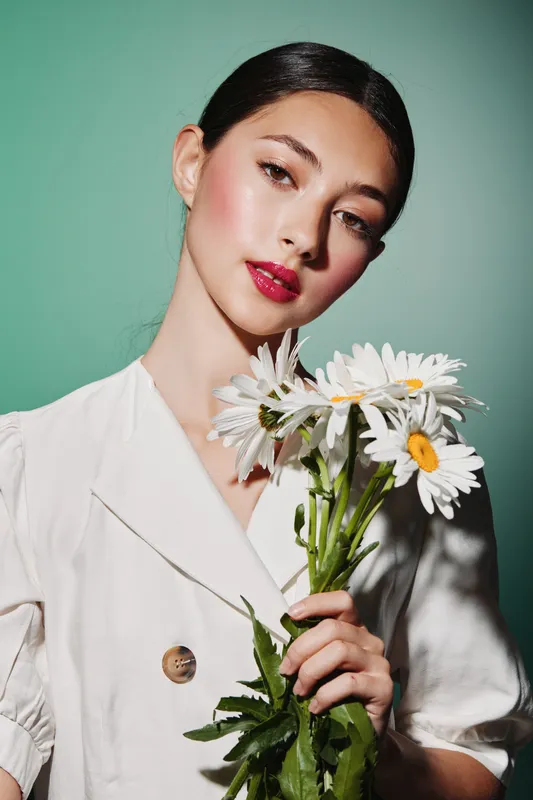 Stylish portrait of a young woman with natural makeup wearing a white outfit and holding a bouquet of daisies against a light green backdrop. Beauty concept for fashion creativity.