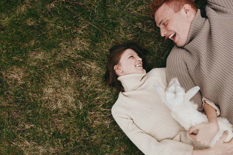 Joyful couple outdoors lying on grass, smiling at each other, embracing a small goat kid, cozy sweaters, sunlit moment, natural tones, relaxed mood, countryside charm with warm light and gentle breeze